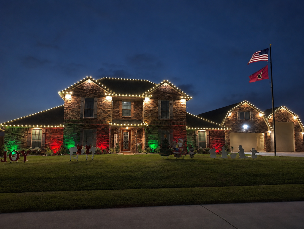 Professional Christmas light installation on a large two-story brick home featuring warm white roofline lights, red and green landscape uplighting, festive yard decorations, and an illuminated American flag at dusk.